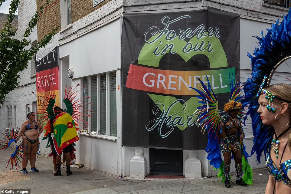 Carnival performers prepare their costumes in the side streets today in front of a poster remembering Grenfell victims